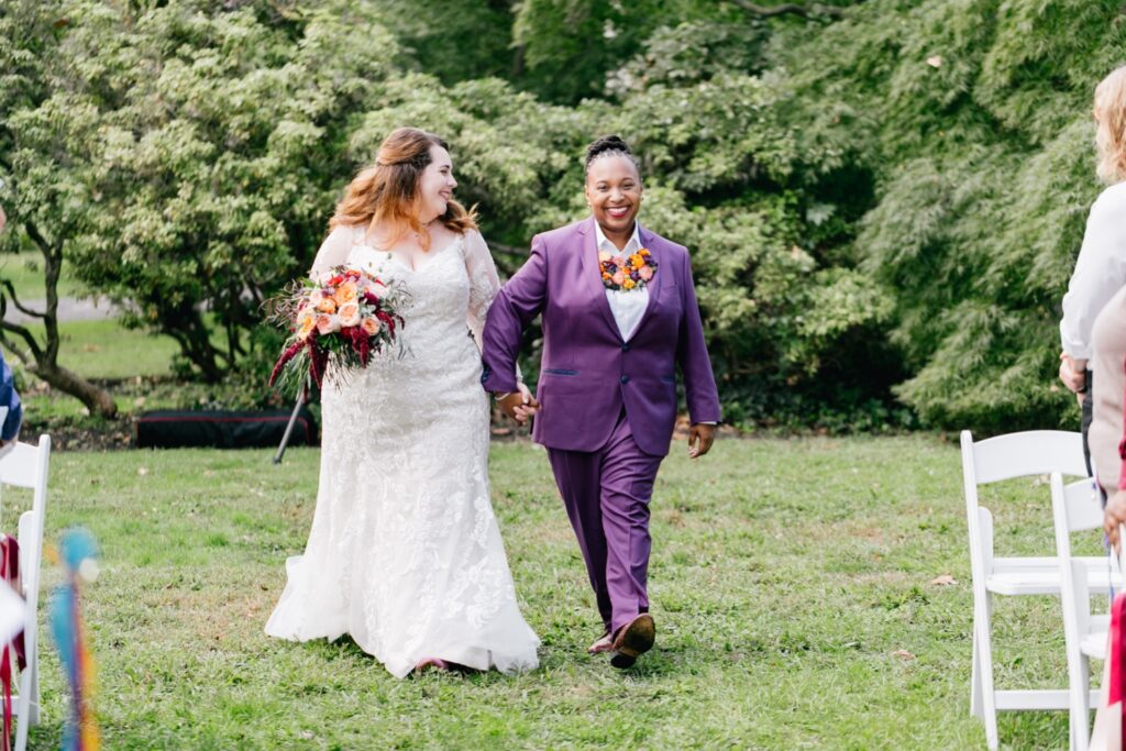 Interracial LGBTQ brides walking down the aisle together during an outdoor wedding ceremony
