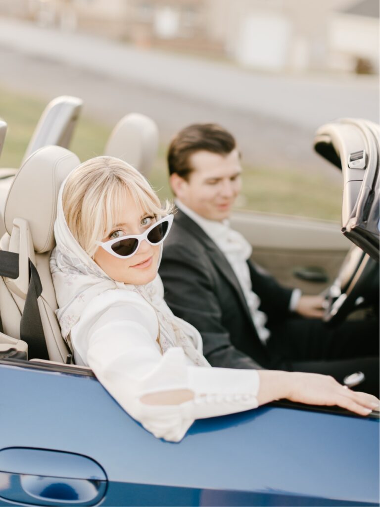 Blonde woman in a classic car with a vintage scarf and sunglasses during a timeless engagement session
