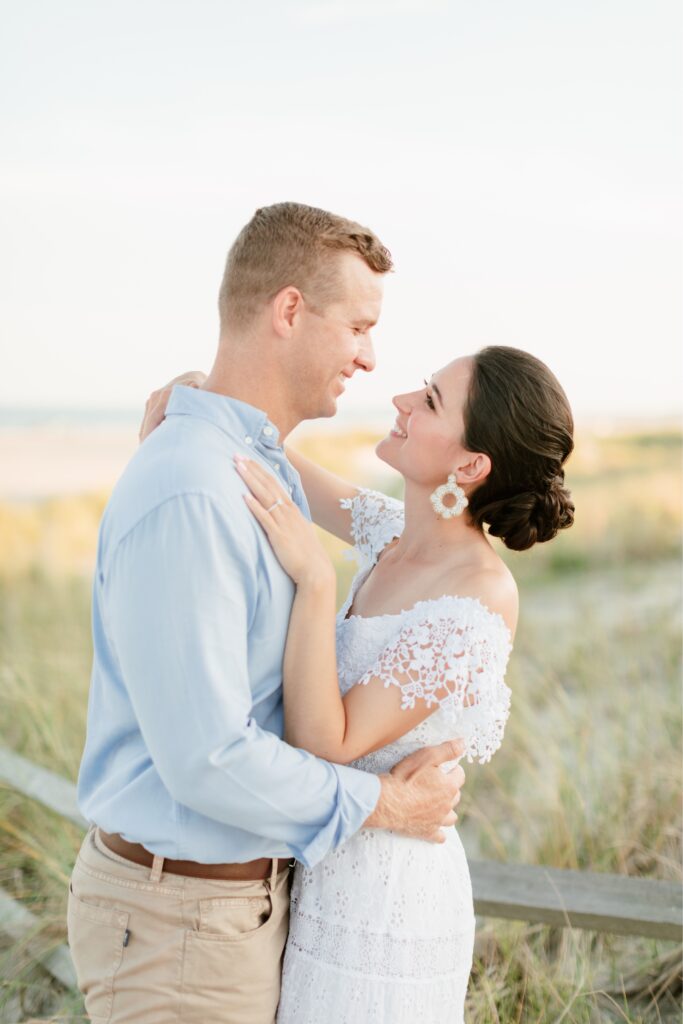Couple smiling at each other on the sand dunes during a beach engagement session by Emily Wren Photography