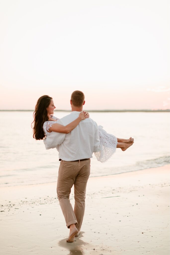 Man carrying his fiancé on the beach during a sunset engagement shoot by Emily Wren Photography