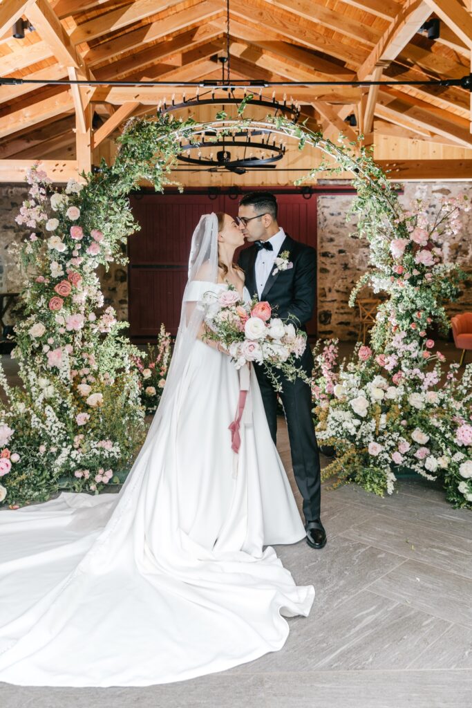 bride and groom kissing in front of floral arch in a barn in Pennsylvania