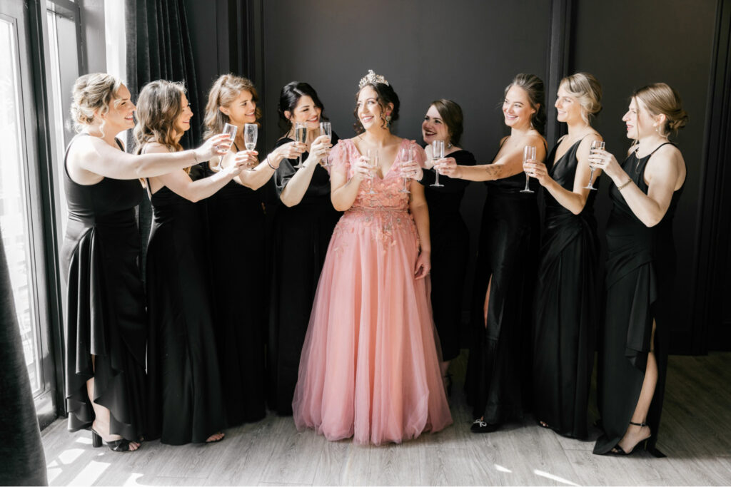 Bride in pink wedding gown drinking champagne with her bridesmaids in black dresses before her Spring wedding in Center City Philadelphia