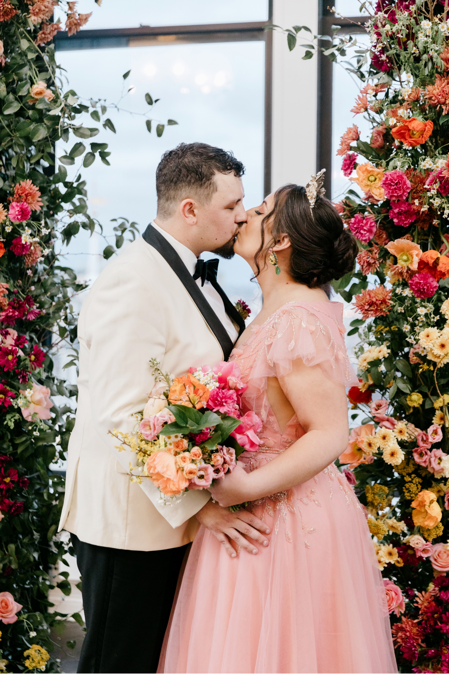 bride and groom standing in front of a colorful floral arch with pink and orange flowers at their wedding ceremony at the Downtown Club