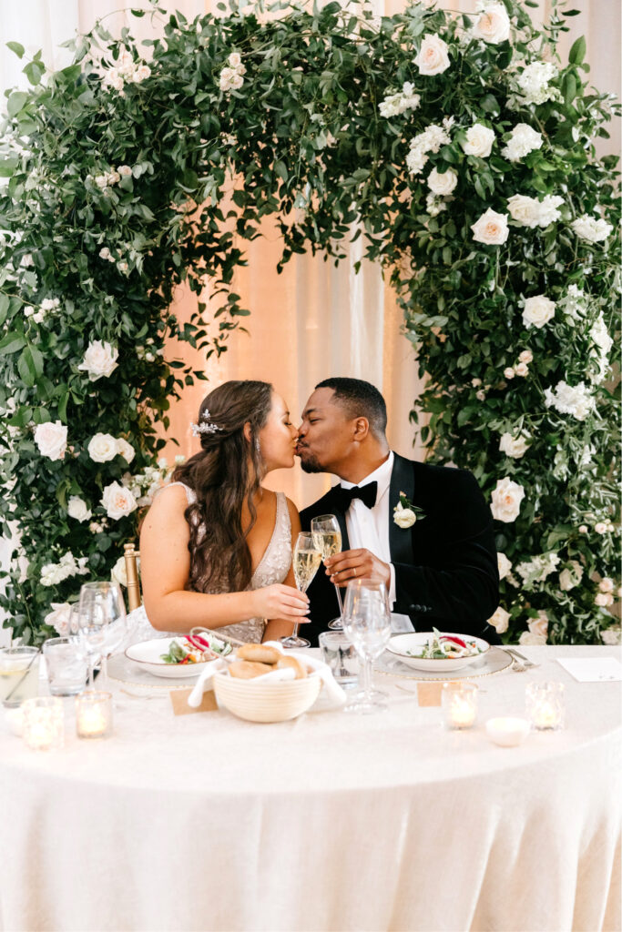 bride & groom at sweetheart table in front of white & pink floral arch