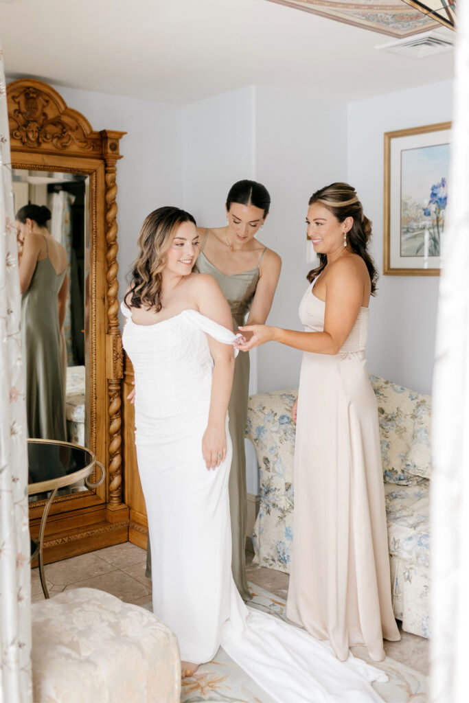 bride getting dressed in her off the shoulder wedding dress with the help of her bridesmaids