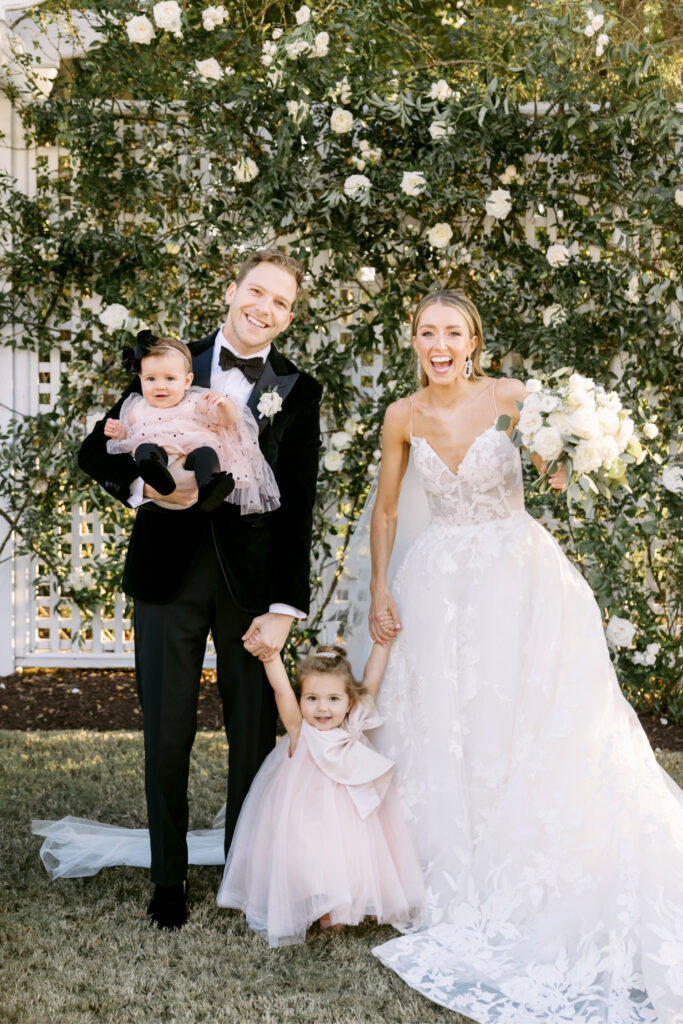 bride & groom with their flower girls in front of greenery and white rose wall