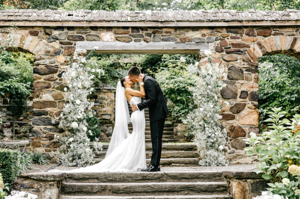 bride & grooms first kiss in front of white babies breath floral wedding arch at Parque Ridley Creek by by Pennsylvania wedding photographer Emily Wren Photography