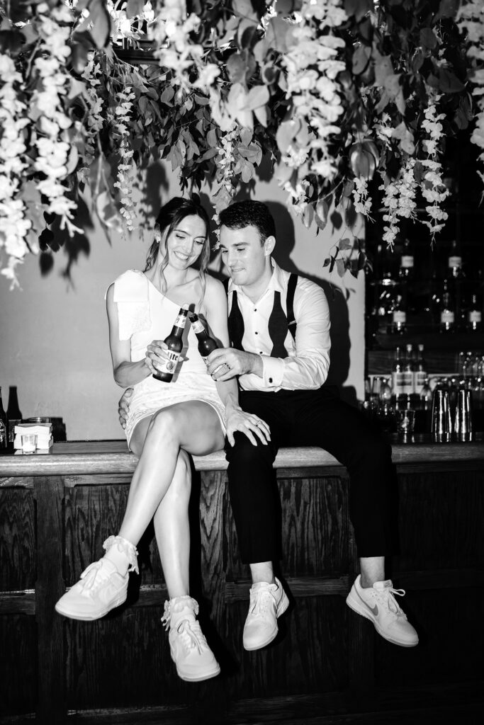 bride and groom sitting on bar drinking a beer by Emily Wren Photography